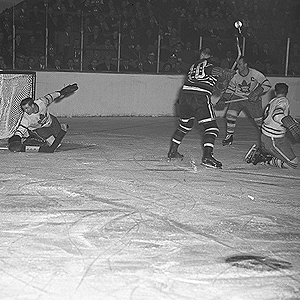 Ice-level during action of a hockey game. A goal-tender reaches down with his blocker to stop a puck. An opposition forward and two of his own players are in front of his net.
