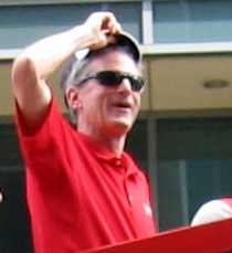A white man in a red polo shirt and sunglasses is standing atop a red double-decker bus, raising his hat, and looking towards the photographer's right.