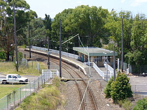 Single-track railway line with overhead electrical catenaries running through a station with a curved platform, a small single-storey flat-roofed station building, and a pedestrian level crossing
