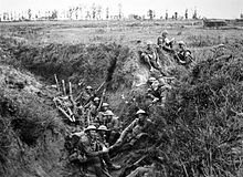 Soldiers wearing helmets and carrying weapons sit in a trench