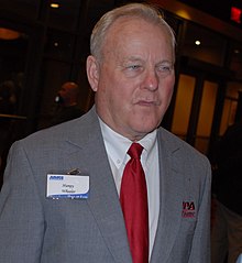 A headshot of NASCAR businessman Humpy Wheeler. Wheeler is displayed wearing a grey suit and red tie.