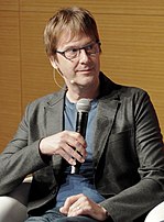 Photograph of Mark Cerny, a middle-aged American man holding a microphone wearing glasses, a blue t-shirt, and a black coat.
