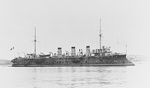 A dark gray ship with a long, forward sloping bow sits idle in the water