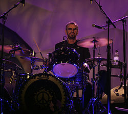 A colour photograph of Ringo Starr playing a dark coloured drum kit on a stage. The background is yellow.