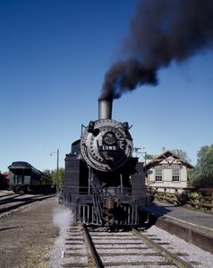 A steam locomotive sitting next to a small passenger station with a vintage passenger coach in the background