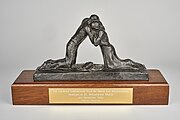 Photograph of The Coventry International Prize for Peace and Reconciliation awarded to Dr Madeleine Sharp in 2004. The award is a metal sculpture of two figures embracing, mounted onto a wooden block with a gold-coloured engraved plaque on the front bearing the inscription "The Coventry International Prize for Peace and Reconciliation Awarded to Dr Madeleine Sharp 14th November 2004 Father Forgive".