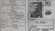 A portion of a small two-page document. On the left is a table with German field names in a Germanic typeface, filled in with handwriting giving names, places and dates. On the right is a black and white photograph of a dark-haired man in a striped top seen from the chest up with a number on the left side at bottom. Next to it are two fingerprints, and below his signature and the date 16 February 1946. Around this are several stamps.