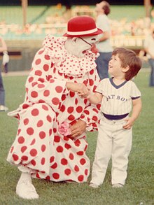 A clown in a white suit with red dots kneels down next to a child