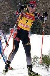 A woman cross country skis uphill towards the camera, holding a red ski pole in each hand. She wears black winter sportswear, a red cap and a yellow jersey with the number 7. A second skier behind her can be seen on the left.