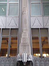 Detail to a building clad in decorative, zig-zagging metal designs, with a single limestone pier rising up in the center of the image, featuring a scale model of the setback skyscraper.