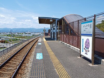 A view of the station platform and track. There is a picture of the station eel mascot character under the station name board.