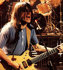 Malcolm, aged 37, plays his guitar with both hands, while singing into a microphone on a stand. His over shoulder-length hair partly obscures his face.