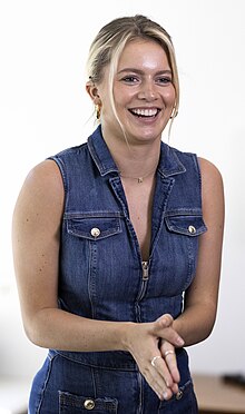 A smiling blonde woman in a sleeveless jean jacket and a visible cochlear implant.