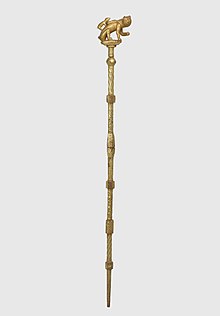 A gold staff topped with a sculpture of a cat and a mouse attempting to crawl inside the cat's bag.