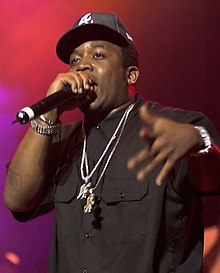 A black male wearing a black shirt, black baseball cap, and two neckchains, speaks into a microphone
