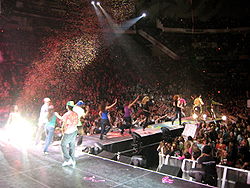 A group of background dancers with colorful clothing running down a catwalk stage as confetti falls from above. At the very end of the catwalk, near a crowd, is a brunette teenager wearing a pink ensemble.