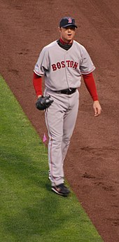 A man in a grey baseball uniform with the word "BOSTON" written across the chest in red letters walks along a baseball field.