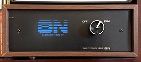 A front view of a wooden box with wood-veneer exterior and a black front. A blue "ON Subscription TV" logo, with the ON letters in a linear gradient, graces the front. A silver knob selects between two options: OFF and ON (stylized like the logo). Beneath the knob is the instruction "TUNE TV TO CH 3 FOR ON".
