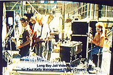 Thirty year old Kelly is shown in left profile playing a guitar in front of a microphone. To his right are two guitarists and a keyboardist amid musical equipment. Behind the keyboard is the drummer at his kit, partly obscured by another microphone. In the background are high fences.