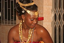 An Akan woman with gold ornaments in her hair, layered gold necklaces, and gold earrings.