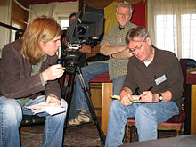 A photograph of four men in a room, three of them sitting down, all surrounding a black videocamera on a black tripod with windows in the background
