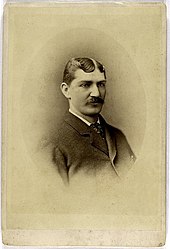 A shoulders-up photograph of a dark-haired man with a thick mustache. He is wearing a dark suit with a dark-patterned tie.
