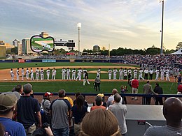 Baseball players in white uniforms with black caps line up along the third base line as they are introduced to the crowd at the ballpark.