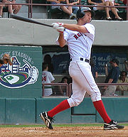 A man in a white baseball uniform and red socks swings left-handed at a baseball.