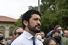 Casar appears with well-kept hair slicked back, a carefully maintained beard and mustache, and dark brown eyes as he looks intently towards the viewer's right, in the direction of the protest leaders. It is clear he is listening intently to the speaker's every word. Soon after this photo was taken, he will speak to the crowd as well. In the background are pro-Palestine and pro-Israel demonstrators, who attempt to yell over one another to communicate their disparate views. Many pro-Palestine demonstrators sport masks and sunglasses to hide their identities.