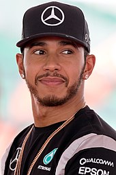 A man in his early thirties sporting a beard and looking to the left of the camera. He is wearing a black baseball cap, a black T-shirt with silver stripes adorning the sides of his sleeves and is sporting a chain necklace around his neck