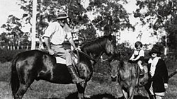 A man on a horse at left looking at camera with a small boy on another horse and a woman standing next to the boy