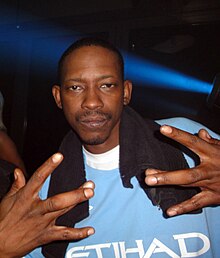 An image of a man wearing blue shirt and flashing a hand sign to the camera.