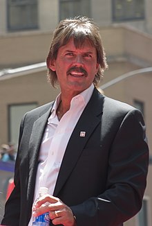Dennis Eckersley in a dark suit with the top of a water bottle in his right hand