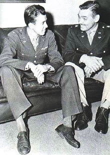 Two men, actor James Stewart and Gable are in their dress uniforms and are seated comfortably on a couch, smiling happily at each other.