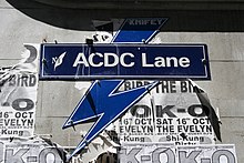 All caps "ACDC" (no dash, no space) followed by "Lane" are seen in off-white lettering on midnight blue background. They are preceded by an off-white stylised M and a feather. Above and below the sign is a mid-blue lightning bolt bordered in white. Both are attached to a brick wall with visible, security head screws. The wall has partly torn, unrelated promotional posters.