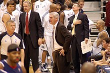 Sloan in a suit, surrounded by several basketball players and a few other coaches, also in suits