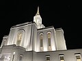 The Orem Utah Temple, seen from below, looking skywards, at night. White parapets stretch tall into the sky, the building has stained glass windows featuring cherry trees. Decorative stonework surrounds the windows with vegetal elements of cherry trees, along with similarly decorated elements along the frieze.