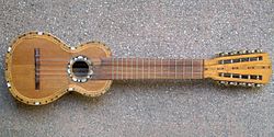 A charango, a small stringed instrument resembling a guitar, lies on a textured surface. The charango features a detailed, decorative inlay around the soundhole and along the edges, with a short neck and multiple tuning pegs.