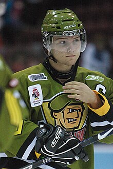 A frontal view of a white, teenage ice hockey player during a game. He is wearing a green, visored helmet and an olive green jersey with a logo consisting of an animated face of a soldier. He appears calm while looking to the right and is reaching for his face with his right hand.