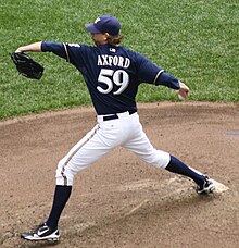 A man wearing a navy blue Brewers jersey, white pants, and a navy blue cap shown in the midst of throwing a ball from the pitcher's mound