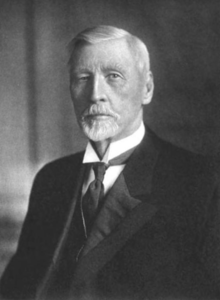 Black and white portrait at bust length of an older man with white hair and a moustache and goatee, wearing a three piece suit and tie.