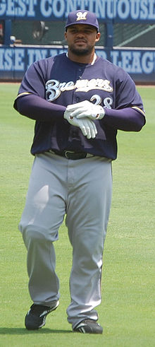 A man wearing a navy blue Brewers jersey, gray pants, and a navy blue cap adjusts his white batting gloves