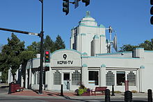 An Art Deco building painted white with turquoise accents. It sits on a corner. Above the door is a sign with a stylized 9 and the call sign KCFW-TV.