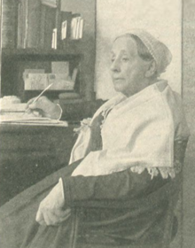 An older white woman, seated; she is wearing a head covering and a white shawl over a dark garment; one arm is resting on a desk with a quill pen in hand
