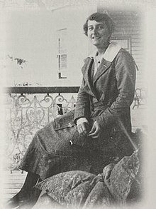 A smiling white woman with short dark hair, seated outdoors, wearing a tweedy skirt and jacket