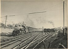 A view of a rail yard adjacent to mainline tracks. A steam locomotive pulls a passenger train on the main line heading towards the photographer, while in the yard tracks another locomotive is seen switching cars. Several other yard tracks are occupied with freight cars. Telltales are visible in the foreground above the tracks.