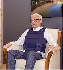 Man with white hair and dark rimmed glasses wears a white dress shirt, dark dress pants, and a navy blue bullet resistant vest while sitting in a beige chair with light wood trim