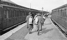 Soldiers walk down a station platform, while on either side of the platform are two trains. The men are wearing slouch hats and are carrying bags over their shoulders.