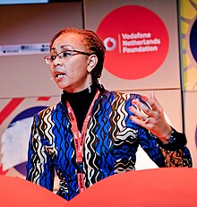 A Black woman in glasses in a blue and black geometric print blouse with her left hand raised