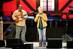 A blonde woman wearing a tan jacket and blue pants, singing into a microphone on a stage. A man playing a guitar is visible in the background.